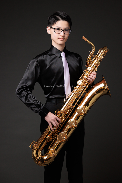 管樂團的名佑這天和同學們一起來拍音樂會要使用的相片，一進門我們就注意到他的低音薩克斯風，瘦高的名佑在鏡頭前很自在啊，這張是名佑站著手拿著他的低音薩克斯風很沈穩地看著鏡頭的相片！