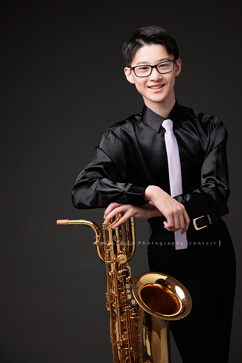 管樂團的名佑這天和同學們一起來拍音樂會要使用的相片，一進門我們就注意到他的低音薩克斯風，瘦高的名佑在鏡頭前很自在啊，這張是名佑站著手扶著他的低音薩克斯風很燦爛地笑著看著鏡頭的相片！