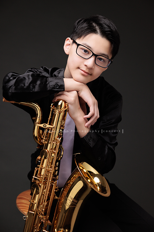 管樂團的名佑這天和同學們一起來拍音樂會要使用的相片，一進門我們就注意到他的低音薩克斯風，瘦高的名佑在鏡頭前很自在啊，這張是名佑坐著手扶著他的低音薩克斯風很有自信地看著鏡頭的相片！