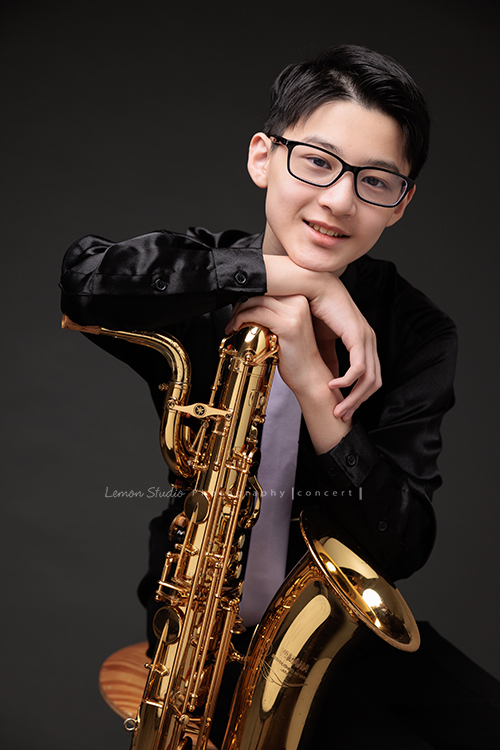 管樂團的名佑這天和同學們一起來拍音樂會要使用的相片，一進門我們就注意到他的低音薩克斯風，瘦高的名佑在鏡頭前很自在啊，這張是名佑坐著手扶著他的低音薩克斯風很有自信地看著鏡頭微笑的相片！