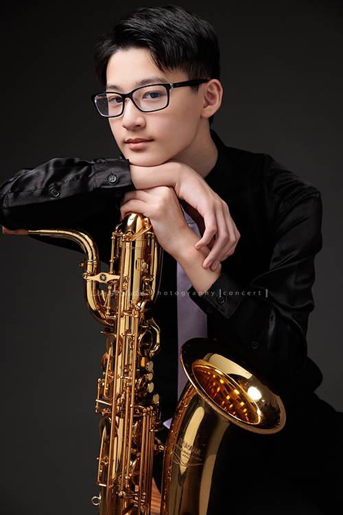 管樂團的名佑這天和同學們一起來拍音樂會要使用的相片，一進門我們就注意到他的低音薩克斯風，瘦高的名佑在鏡頭前很自在啊，這張是名佑坐著手扶著他的低音薩克斯風很有藝術氣息、名佑看著鏡頭的相片！