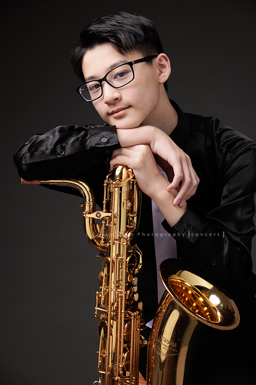 管樂團的名佑這天和同學們一起來拍音樂會要使用的相片，一進門我們就注意到他的低音薩克斯風，瘦高的名佑在鏡頭前很自在啊，這張是名佑坐著手扶著他的低音薩克斯風很有藝術氣息、名佑微笑地看著鏡頭的相片！
