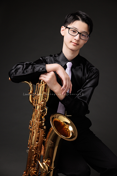 管樂團的名佑這天和同學們一起來拍音樂會要使用的相片，一進門我們就注意到他的低音薩克斯風，瘦高的名佑在鏡頭前很自在啊，這張是名佑坐著手扶著他的低音薩克斯風很有架勢地看著鏡頭的相片