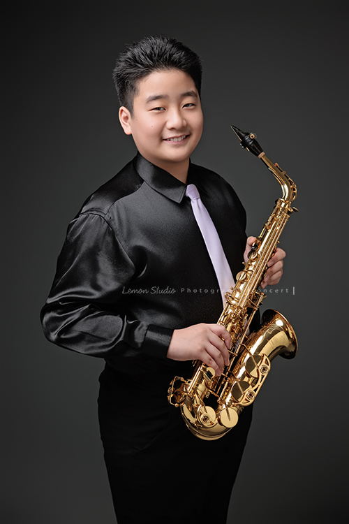 在同一個管樂團的軒＆宸兄弟檔來拍照，吹奏薩克斯風開朗的兩人，在鏡頭前很輕鬆自在地做自己，這張是弟弟宸站著著拿著薩克斯風很有自信地笑著且看著鏡頭的一張很帥氣的相片！