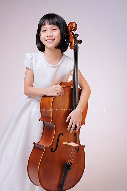 芯參加學校的弦樂團正在學習大提琴，為了即將來到的音樂會演出來拍音樂會寫真，雖然一開始拍照時有點小緊張，但逐漸地在鏡頭前越來越自在，結果捕捉到許多芯好可愛的表情及畫面，這張是芯站在大提琴旁、手扶著大提琴沒有看鏡頭微笑著的一張音樂會寫真照！