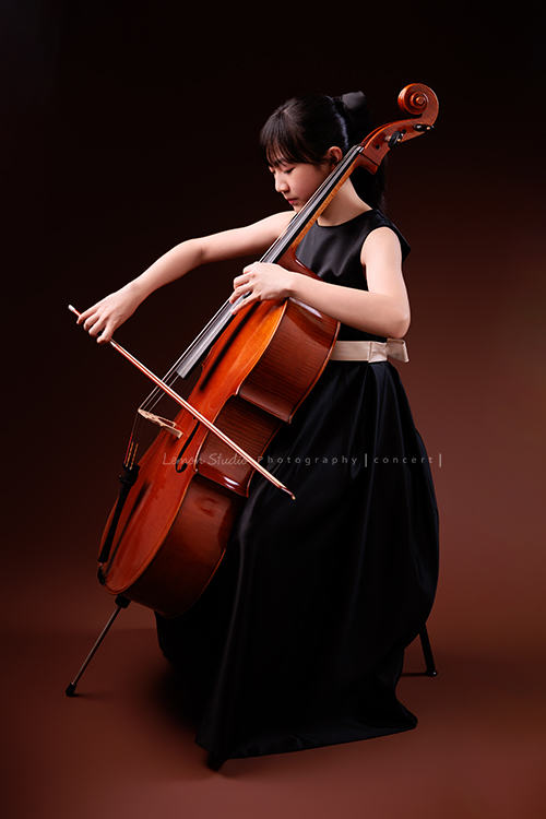 Lucie為了即將來到的音樂會帶著大提琴來拍音樂會寫真照，很有氣質的Lucie，笑起來很甜美，這張是Lucie坐在椅子上，正非常專注地拉著大提琴的側面的全身照！