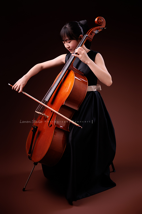 Lucie為了即將來到的音樂會帶著大提琴來拍音樂會寫真照，很有氣質的Lucie，笑起來很甜美，這張是Lucie坐在椅子上，帶著微笑正非常專注地拉著大提琴的側面的全身照！