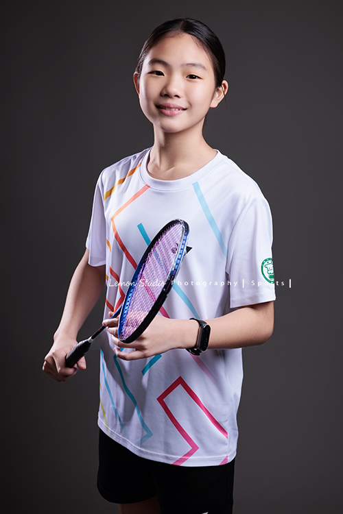 這次國小羽球隊請我們拍球隊的運動寫真照，運動的孩子們有一種很專注內斂的氣質，這次還有拍孩子們打球的瞬間，好有力量好美啊，這張是羽球隊裡一位女孩的運動寫真照，女孩兩手拿球拍、看著鏡頭很甜美地微笑！