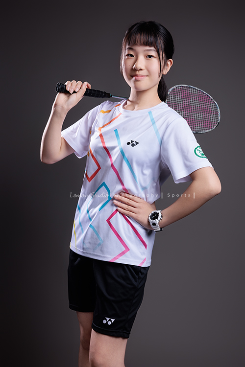 這次國小羽球隊請我們拍球隊的運動寫真照，運動的孩子們有一種很專注內斂的氣質，這次還有拍孩子們打球的瞬間，好有力量好美啊，這張是羽球隊裡一位女孩的運動寫真照，女孩一手拿著球拍放在肩膀後側，一手叉腰，帶著自信微笑地看著鏡頭！