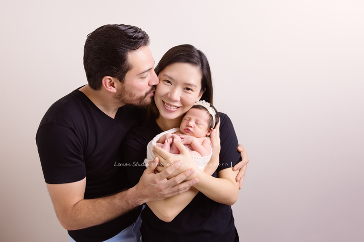 Diana媽麻和先生帶著剛出生的寶寶來拍新生兒攝影，外國人的爸拔和媽麻一起抱著寶寶，三人之間的互動自然又真摯，這是他們第一次的全家福，也是送給寶寶最有溫度美好的出生禮物，這張是爸拔媽麻將寶寶抱在他們兩人的前方，兩人充滿愛地抱著寶寶、爸拔親著媽麻的臉頰、媽麻看著鏡頭微笑的幸福模樣！
