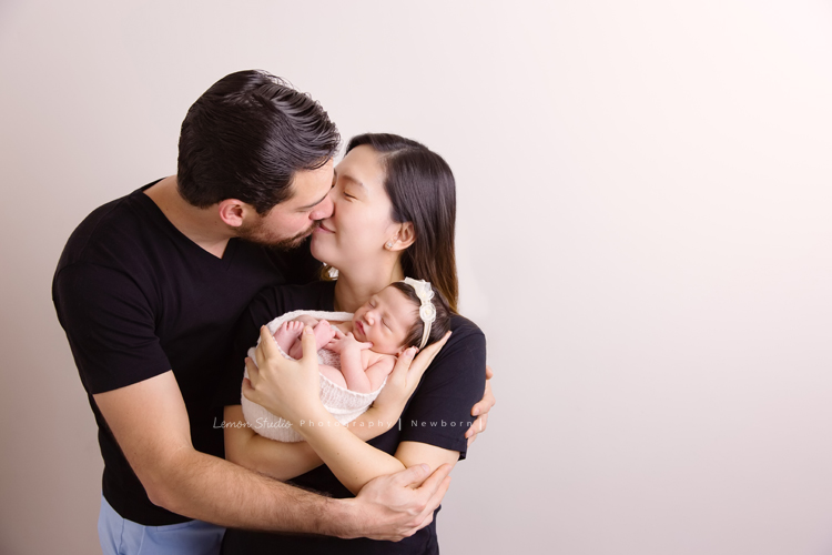 Diana媽麻和先生帶著剛出生的寶寶來拍新生兒攝影，外國人的爸拔和媽麻一起抱著寶寶，三人之間的互動自然又真摯，這是他們第一次的全家福，也是送給寶寶最有溫度美好的出生禮物，這張是爸拔媽麻將寶寶抱在他們兩人的前方，爸拔媽麻兩人親吻著並充滿愛地抱著寶寶的幸福模樣！