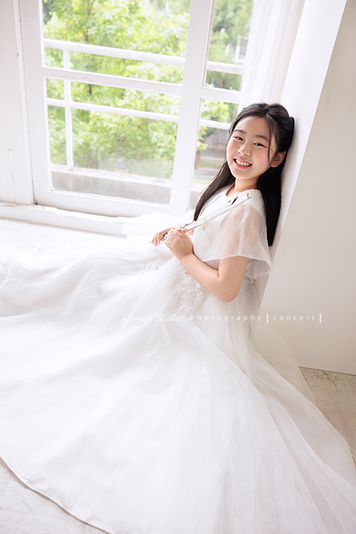 宥欣自小喜愛長笛，樂於在台上演出並與大家分享美好的音樂，9歲時即以非凡的音樂表現脫穎而出，成為台灣歷年來長笛組最年輕首位榮獲日本第33回古典音樂大賽全國大會總決賽長笛部國小中年級最高名次，今年剛滿11歲的她，將於11月底舉辦人生中的第一場獨奏會，有興趣的人可以留言跟我們索票喔，這張是宥欣穿著白色禮服雙手拿著長笛，坐在地上靠著牆，看著鏡頭笑得很甜美的一張長笛音樂寫真！
