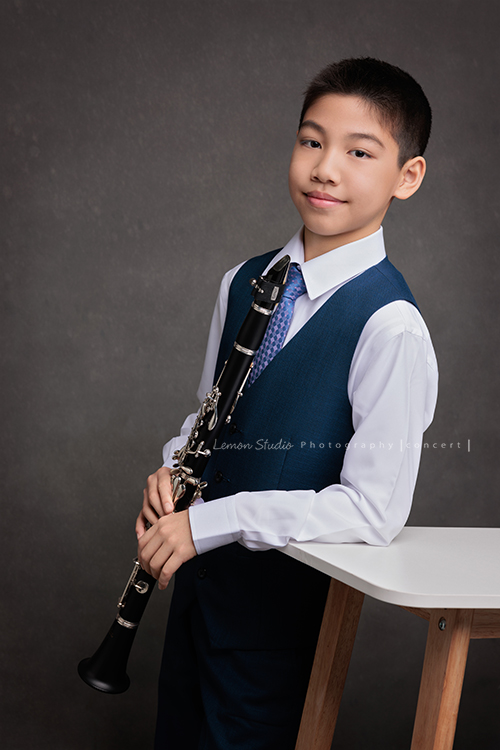 圻和同學們即將舉辦一場音樂會，身為管樂團的一員圻吹奏的是豎笛，換上西裝的圻多了份帥氣與沉穩，鏡頭裡的他自信而有氣質，這張是圻一手拿著豎笛、另一手手肘靠在桌面上、看著鏡頭微微地笑的音樂會相片！