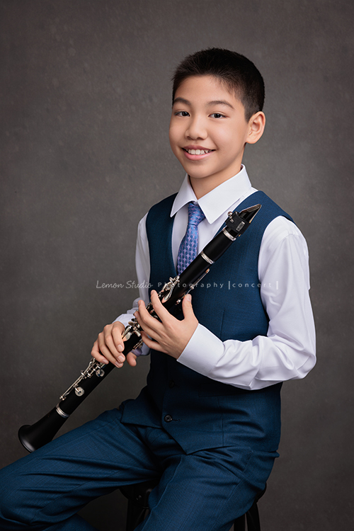 圻和同學們即將舉辦一場音樂會，身為管樂團的一員圻吹奏的是豎笛，換上西裝的圻多了份帥氣與沉穩，鏡頭裡的他自信而有氣質，這張是圻兩手拿著豎笛坐在椅子上看著鏡頭微笑的音樂會相片！