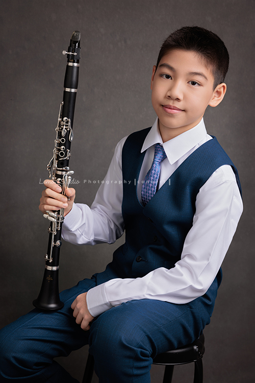 圻和同學們即將舉辦一場音樂會，身為管樂團的一員圻吹奏的是豎笛，換上西裝的圻多了份帥氣與沉穩，鏡頭裡的他自信而有氣質，這張是圻一手拿著豎笛坐在椅子上、另一手放在腳上，很沈穩地看著鏡頭的音樂會相片！