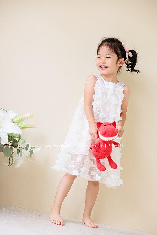 Jess媽一家第三次來拍照，這次是為了紀念雅滿四歲的時光，一家人總是自然又自在，照片裡流露出的，是日常中最真實舒服的模樣，這張是雅穿白色洋裝、手拿著一隻紅色的娃娃，看著窗外笑得很可愛的模樣！