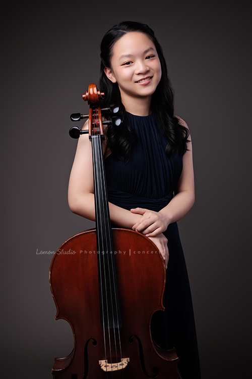 Sophia 帶著她的大提琴來拍照，深藍禮服如湖水靜靜流動，淡妝與盤起的髮絲勾勒出沉靜優雅，微笑時彷彿音符也跟著亮了起來，這張是Sophia站著雙手扶在大提琴上看著鏡頭微笑的模樣！