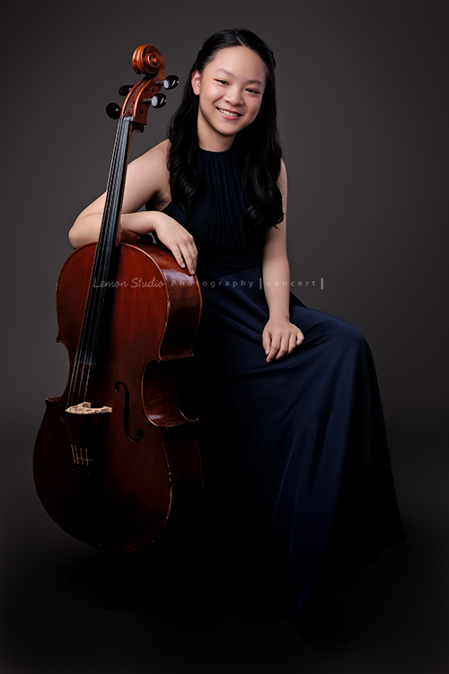 Sophia 帶著她的大提琴來拍照，深藍禮服如湖水靜靜流動，淡妝與盤起的髮絲勾勒出沉靜優雅，微笑時彷彿音符也跟著亮了起來，這張是Sophia很優雅地坐在椅子上，一手扶著大提琴看著鏡頭笑得很甜美的模樣！