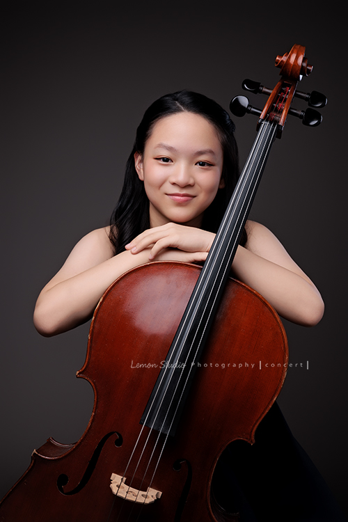 Sophia 帶著她的大提琴來拍照，深藍禮服如湖水靜靜流動，淡妝與盤起的髮絲勾勒出沉靜優雅，微笑時彷彿音符也跟著亮了起來，這張是Sophia坐著雙手扶在大提琴上看著鏡頭微笑的模樣！