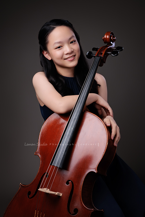 Sophia 帶著她的大提琴來拍照，深藍禮服如湖水靜靜流動，淡妝與盤起的髮絲勾勒出沉靜優雅，微笑時彷彿音符也跟著亮了起來，這張是Sophia坐著雙手扶在大提琴上看著鏡頭笑得很甜美的模樣！