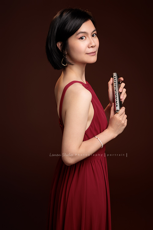 知名口琴演奏家衍華，近日為音樂形象照拍攝，緩緩展開她與音樂的對話，歷經無數舞台洗禮，屢獲殊榮並擔任評審的她，舉手投足間皆是樂音流動的痕跡，彷彿音符化形，翩然於鏡頭之中，這張是衍華身穿紅色的禮服手拿著口琴，側身轉過頭來沒有看鏡頭微微地笑的一張很美的音樂形象照！