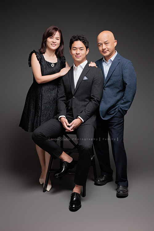 Cindy 媽麻一家來拍照，在美國念書的兒子瑋，帥氣又溫暖，一家人換上正式與休閒的服裝，每一張照片都記錄下最自然的互動與情感，這張是兒子瑋穿著西裝帥氣地坐在椅子上、爸爸和媽媽站在他的兩邊，一家三口很有質感及自然的全家福寫真！