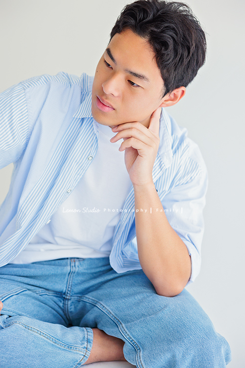 Cindy 媽麻一家來拍照，在美國念書的兒子瑋，帥氣又溫暖，一家人換上正式與休閒的服裝，每一張照片都記錄下最自然的互動與情感，這張是兒子瑋坐在白色箱子上，穿著藍色系的休閒服飾，一手托著頭，臉側向一邊沒有看鏡頭，是一張很帥氣的個人寫真！