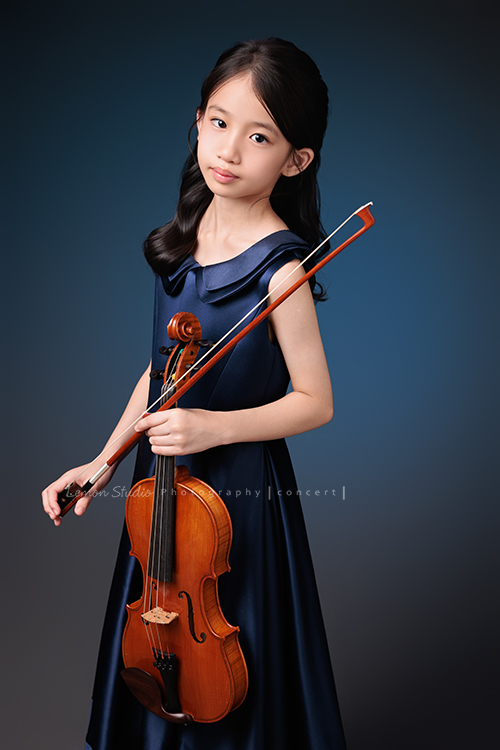 Yvonne 帶著小提琴走進鏡頭，深藍小禮服襯出她的氣質與靜謐，簡單又典雅，她不特意擺姿勢，每一張都自然又有味道，這張是Yvonne站著一手拿著小提琴在身體前方、一手拿弓，很自然地看著鏡頭的音樂寫真照！
