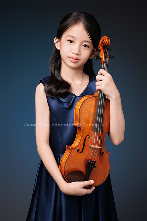 Yvonne 帶著小提琴走進鏡頭，深藍小禮服襯出她的氣質與靜謐，簡單又典雅，她不特意擺姿勢，每一張都自然又有味道，這張是Yvonne站著兩手拿著小提琴在身體前方，很自然地看著鏡頭的音樂寫真照！