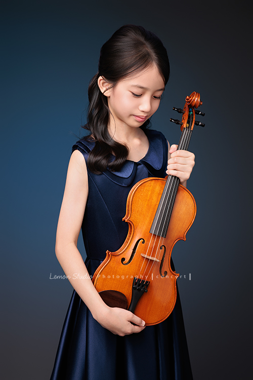 Yvonne 帶著小提琴走進鏡頭，深藍小禮服襯出她的氣質與靜謐，簡單又典雅，她不特意擺姿勢，每一張都自然又有味道，這張是Yvonne站著兩手拿著小提琴在身體前方，低頭沒有看鏡頭的音樂寫真照！