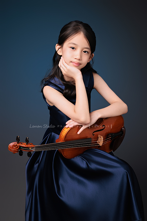 Yvonne 帶著小提琴走進鏡頭，深藍小禮服襯出她的氣質與靜謐，簡單又典雅，她不特意擺姿勢，每一張都自然又有味道，這張是Yvonne坐在椅子上一手托著頭、一手扶著小提琴，看著鏡頭微笑的音樂寫真照！