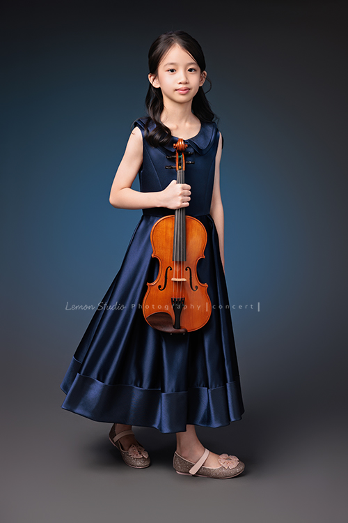 Yvonne 帶著小提琴走進鏡頭，深藍小禮服襯出她的氣質與靜謐，簡單又典雅，她不特意擺姿勢，每一張都自然又有味道，這張是Yvonne站著一手拿著小提琴在身體前方，很自然地看著鏡頭微笑的音樂寫真照！