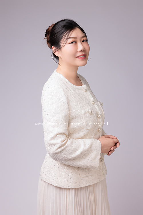 音樂治療師婷婷來拍照,拍完一套正式服裝的形象照後,她換上白色洋裝,輕輕抱著吉他入鏡,這張是音樂治療師婷婷穿著正式的一套白色服裝,兩手交叉在身體前方、側身頭轉過來看著鏡頭微笑的一張形象照!