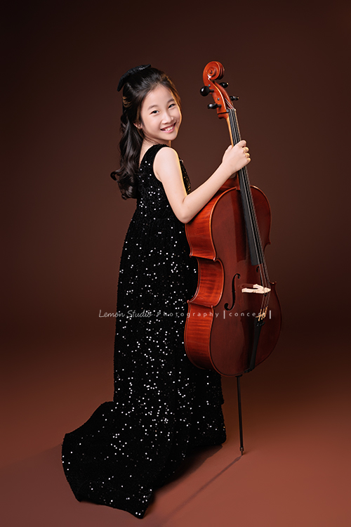 Tristan & Natalie兄妹倆帶著大提琴來拍照，和同學們一起為音樂會做準備，兄妹倆活潑又有默契，這次大家準備的團服也格外亮眼，每一張照片都展現出屬於他們自己的風格與魅力，這張是Natalie背對鏡頭站在大提琴旁、一手拿著大提琴，Natalie轉頭看著鏡頭微笑的大提琴音樂寫真照！
