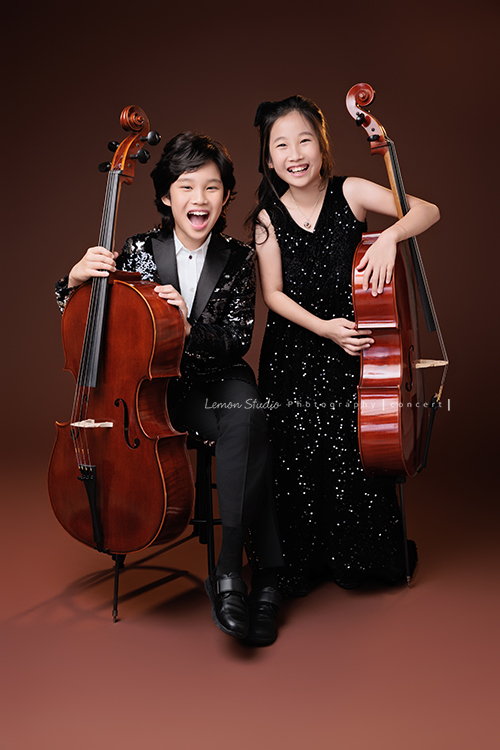 Tristan & Natalie兄妹倆帶著大提琴來拍照，和同學們一起為音樂會做準備，兄妹倆活潑又有默契，這次大家準備的團服也格外亮眼，每一張照片都展現出屬於他們自己的風格與魅力，這張是Tristan坐著Natalie站著，兩人各扶著自己的大提琴」笑得很開心的兩人的大提琴音樂會寫真合照！
