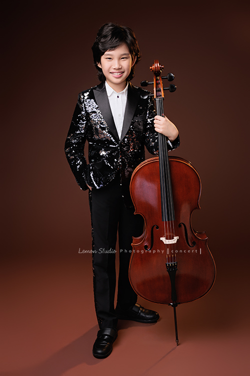 Tristan & Natalie兄妹倆帶著大提琴來拍照，和同學們一起為音樂會做準備，兄妹倆活潑又有默契，這次大家準備的團服也格外亮眼，每一張照片都展現出屬於他們自己的風格與魅力，這張是Tristan站著一手扶著大提琴一手放口袋，Tristan看著鏡頭微笑的大提琴音樂寫真照！