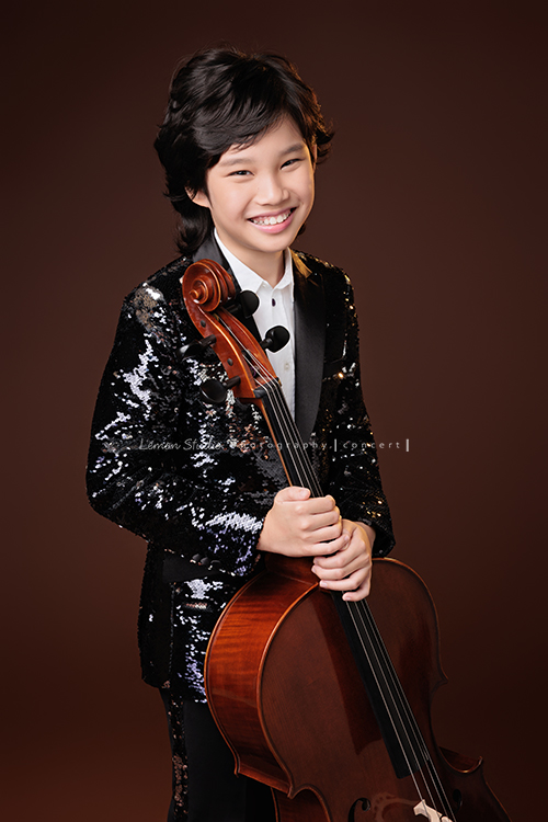 Tristan & Natalie兄妹倆帶著大提琴來拍照，和同學們一起為音樂會做準備，兄妹倆活潑又有默契，這次大家準備的團服也格外亮眼，每一張照片都展現出屬於他們自己的風格與魅力，這張是Tristan站著雙手扶著大提琴，Tristan看著鏡頭微笑的大提琴音樂寫真照！