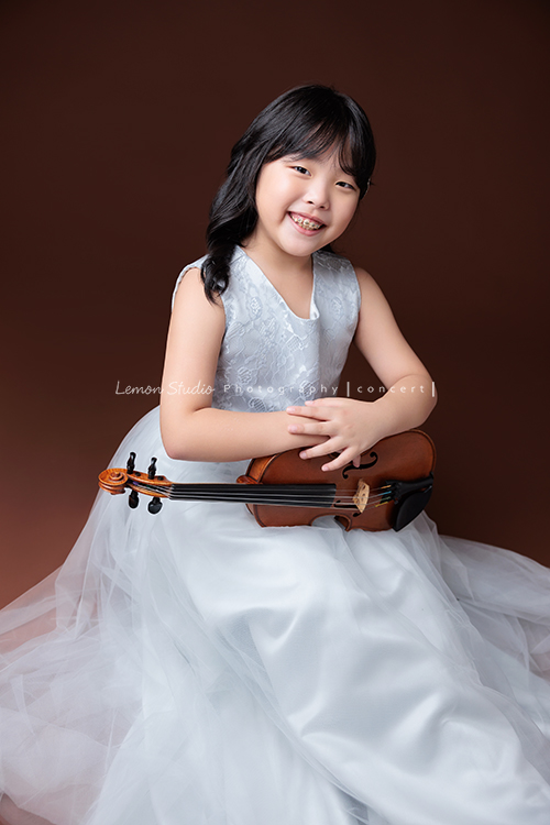 Jonathan & Chloe兄妹倆來拍提琴音樂寫真，再次站在鏡頭前，今年的 Jonathan 明顯長大了許多，Chloe非常活潑可愛，主動幫忙帶動現場氣氛，讓整個拍攝過程溫馨又美好，這張是Chloe坐著裙子擺放地很漂亮、雙手靠在提琴上、看著鏡頭笑得很甜美的一張音樂寫真照！