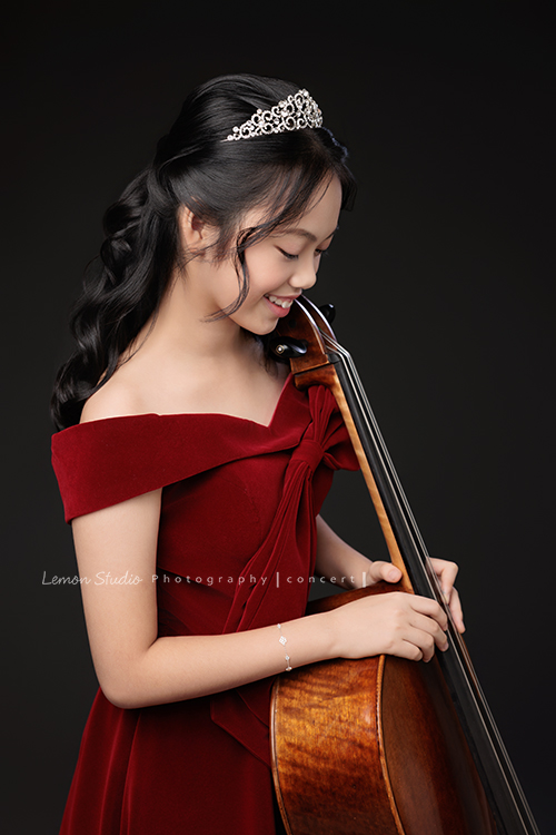 音樂班的Yuko即將畢業，帶著她的大提琴來到鏡頭前，在不同的禮服與不同的樂器之間，看見不一樣的她，這張是Yuko身穿一襲紅色禮服、在紅色背景前、大提琴在身體前方、雙手扶著大提琴、低頭看著大提琴微笑的音樂班畢業音樂寫真！