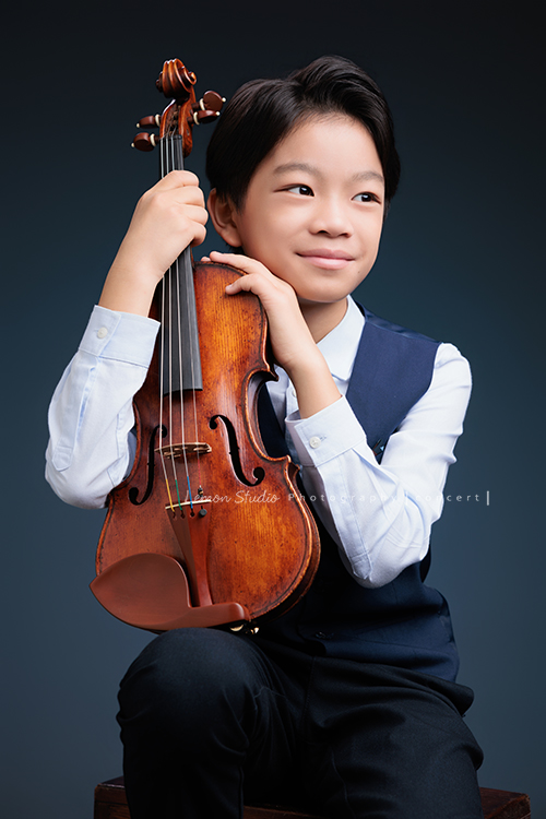 喆即將從國小弦樂團畢業，和同學們一起來拍音樂寫真照，拍照當天在現場很認真地拉琴，我們也不知不覺成了他的粉絲，這張是喆坐著提琴在身體前方、喆雙手扶著提琴沒有看鏡頭看旁邊的遠方處微笑著、很有意境的一張音樂寫真照！