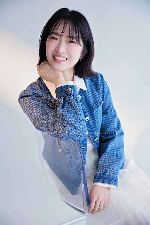 講師Tiffany來拍照，這次我們一起整理出帶點日韓系的風格，有些細微的表情與狀態，很耐看，這張是Tiffany身穿藍色短版外套及白色長裙坐在椅子上、身體靠著椅子、一手放在脖子處、看著鏡頭微笑著的一張形象照！