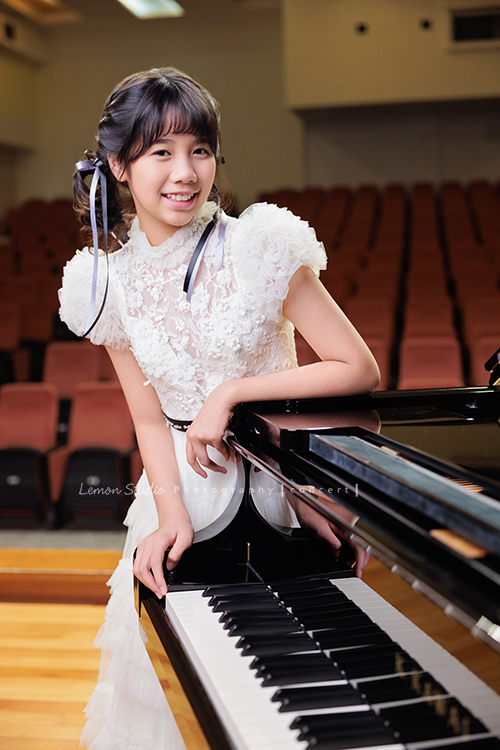 音樂班的 Lily畢業前來拍音樂班畢業音樂寫真照，Lily的活潑與大方在鏡頭前自然流露，這張是Lily穿著一襲可愛的白色禮服、站在鋼琴旁手扶在鋼琴上、看著鏡頭笑得很甜美的一張音樂班畢業音樂寫真照！
