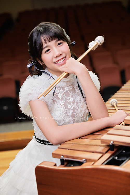 音樂班的 Lily畢業前來拍音樂班畢業音樂寫真照，Lily的活潑與大方在鏡頭前自然流露，這張是Lily穿著一襲可愛的白色禮服、坐在馬林巴木琴旁、一手拿著琴槌在下巴處、一手拿著琴槌放在琴上、看著鏡頭笑得很甜美的一張音樂班畢業音樂寫真照！