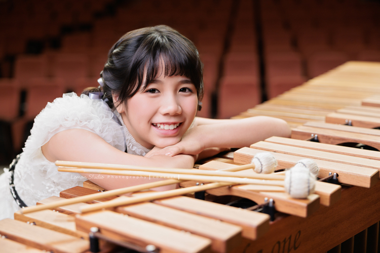 音樂班的 Lily畢業前來拍音樂班畢業音樂寫真照，Lily的活潑與大方在鏡頭前自然流露，這張是Lily穿著一襲可愛的白色禮服、坐在馬林巴木琴旁、琴槌在馬林巴木琴上、Lily趴在琴上、看著鏡頭笑得很甜美的一張音樂班畢業音樂寫真照！