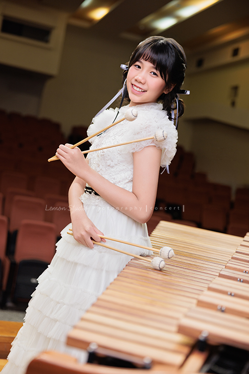 音樂班的 Lily畢業前來拍音樂班畢業音樂寫真照，Lily的活潑與大方在鏡頭前自然流露，這張是Lily穿著一襲可愛的白色禮服、站在馬林巴木琴旁、一手拿著琴槌在身體前方、一手拿著琴槌輕放在琴上、看著鏡頭笑得很甜美的一張音樂班畢業音樂寫真照！